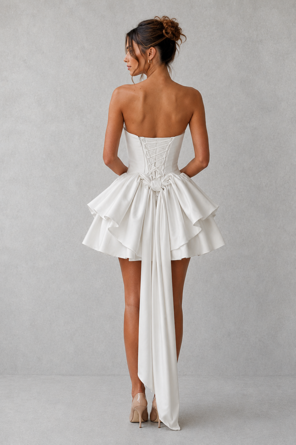 Woman wearing a strapless satin mini dress featuring a softly draped cowl neckline, structured boned bodice, lace-up back, voluminous skirt with pleated godet panels, and a detachable statement bow, ideal for parties, weddings, and special occasions.